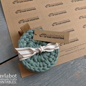 May include: Handmade crocheted car coasters in a teal green colour with a white and tan striped ribbon tied around them. The coasters are on a brown surface with a sheet of printable labels that say "Handmade Car Coasters".