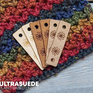 May include: Five Ultrasuede plant-based labels in varying shades of beige and brown, each with a daisy design and a hole at the top. The labels are displayed on a colorful, textured crochet fabric. The text "ULTRASUEDE PLANT-BASED LABELS" is visible.