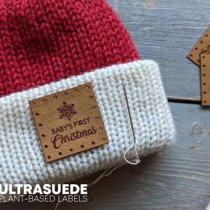 May include: A red and white knit hat featuring a square tan label with the text "BABY'S FIRST Christmas" and a snowflake icon. A silver sewing needle with white thread is positioned next to the label, which is attached to the white brim.