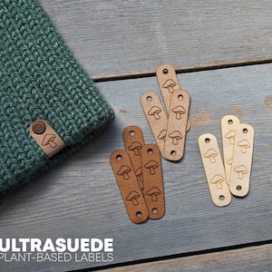 May include: A teal knitted beanie with a leather tag and several leather labels. The labels are tan and brown with a mushroom design. The text "ULTRASUEDE PLANT-BASED LABELS" is visible.