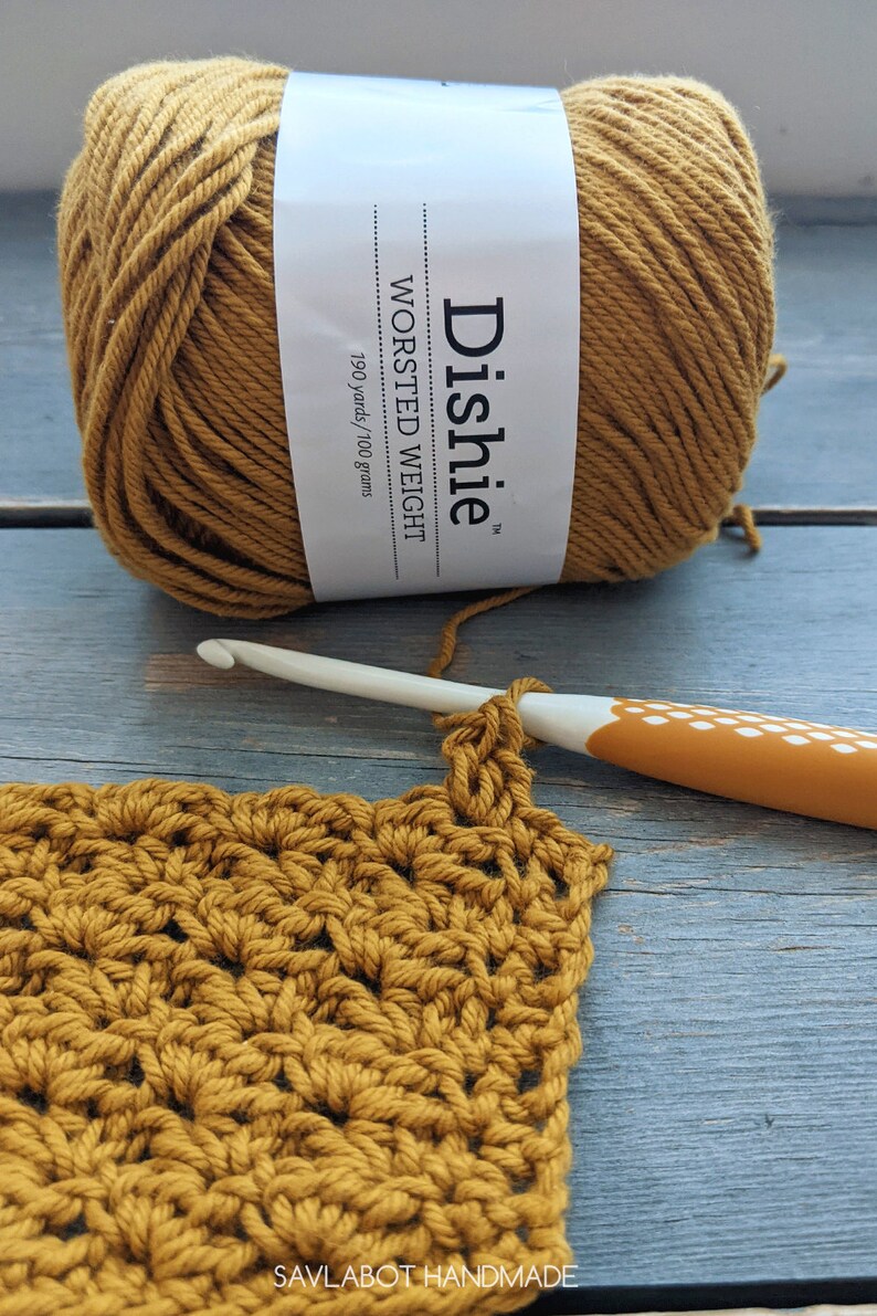 May include: A ball of mustard yellow worsted weight yarn labeled "Dachie" with 190 yards/100 grams. A crochet hook is shown with a crocheted square made with the yarn.