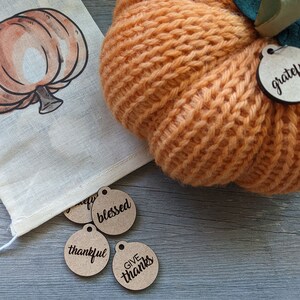 May include: A knitted orange pumpkin with a green leaf and a brown wooden tag that says "grateful". There are four other brown wooden tags with the words "blessed", "thankful", "give", and "thanks" on them.