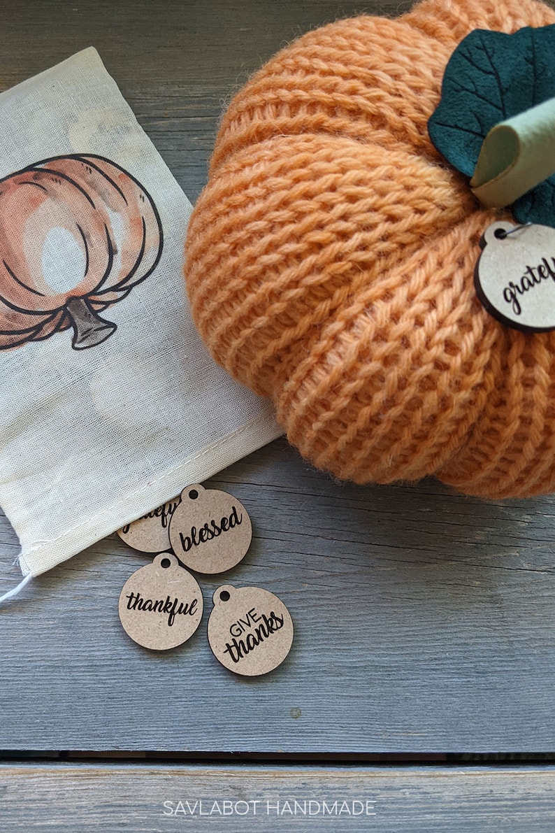 May include: A knitted orange pumpkin with a green leaf and a brown wooden tag that says "grateful". There are four other brown wooden tags with the words "blessed", "thankful", "give", and "thanks" on them.