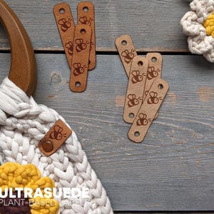 May include: A white macrame bag with a wooden handle and a yellow flower accent, accompanied by several tan rectangular labels featuring a bee graphic and a hole at each end, labeled ULTRASUEDE PLANT-BASED LABELS.