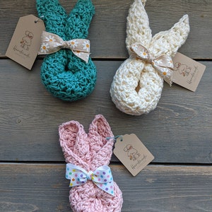 May include: Three crocheted bunny shaped stuffed toys with bows. The bunnies are teal, white, and pink. Each bunny has a brown tag with the text "Handmade with Love" and a small illustration of a bunny.