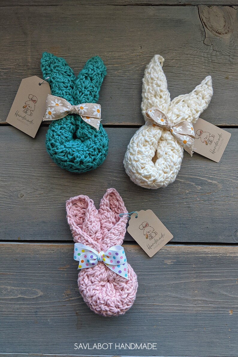 May include: Three crocheted bunny shaped stuffed toys with bows. The bunnies are teal, white, and pink. Each bunny has a brown tag with the text "Handmade with Love" and a small illustration of a bunny.