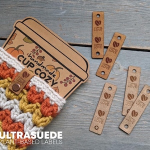 Könnte beinhalten: Ein gehäkelter Tassenwärmer aus weißem, senfgelbem und rostorangem Garn mit einem braunen Lederetikett, das ein Kaffeebohnensymbol und den Text COFFEE LOVE zeigt. Daneben liegen fünf rechteckige braune Lederetiketten mit demselben Kaffeebohnenmotiv.