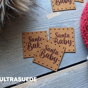 May include: Square tan labels with pre-punched holes along the edges, featuring the text Santa Baby in a dark brown script font. These small, flat, square-shaped tags are designed for attachment to garments or accessories with visible stitching holes.