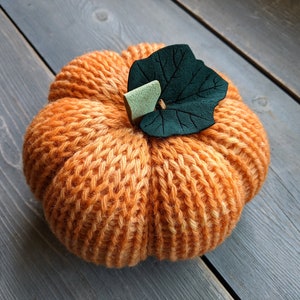 May include: A knitted orange pumpkin with a green felt leaf on top. The pumpkin is sitting on a gray wooden surface.