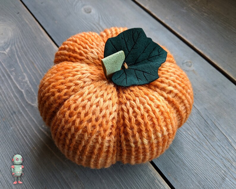 May include: A knitted orange pumpkin with a green felt leaf on top. The pumpkin is sitting on a gray wooden surface.