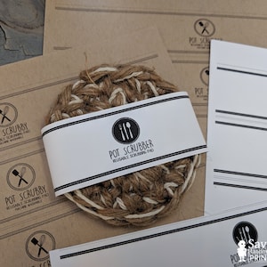 May include: A set of brown paper labels with black text that reads "Pot Scrubby Reusable Scrubbing Pad Machine Washable" and a black and white illustration of a whisk and spatula. The labels are next to a brown and white woven pot scrubber with a white label that reads "Pot Scrubber Reusable Scrubbing Pad".