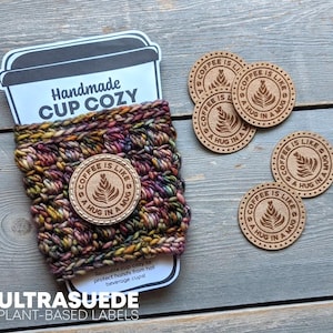 Puede incluir: Una funda para taza de punto grueso multicolor con un parche circular de cuero color canela que incluye el texto "COFFEE IS LIKE A HUG IN A MUG" y un gráfico de hoja. Cuatro parches de cuero circulares a juego se sitúan a la derecha.