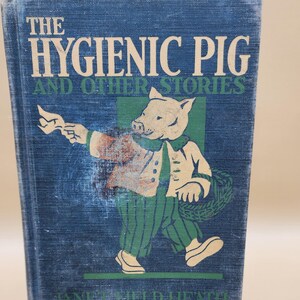May include: A blue book with the title "The Hygienic Pig and Other Stories" by Janet Field Heath. The cover features an illustration of a pig wearing a white shirt, green pants, and a white hat, holding a wreath.