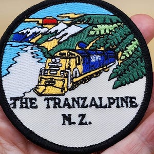May include: A round embroidered patch with a yellow and blue train traveling through a mountain landscape. The text "THE TRANZALPINE N.Z." is below the train.