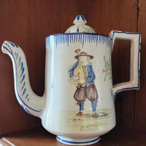 May include: A white ceramic teapot with a blue and white striped design. The teapot features a hand-painted scene of a person playing a bagpipe. The teapot has a lid with a blue knob.