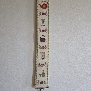 May include: A long, vertical cream-colored fabric wall hanging featuring cross-stitch embroidery. The design includes a red teapot, a blue goblet, a black cauldron, an hourglass, and a green bottle with a glass, separated by small floral motifs.