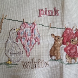 May include: Cross-stitch embroidery featuring a goose, bunnies, and laundry hanging on a clothesline. The words "pink", "white", and "red" are stitched above and below the objects. The color palette includes pink, white, red, brown, and green.