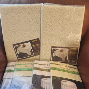May include: A set of five white vinyl tablecloths with lace patterns. The tablecloths are packaged in plastic and labeled with the brand name "Vytex Vinyl Lace" and the size of each tablecloth. The tablecloths are available in 52x72 inch, 70 inch round, and 70 inch square sizes.