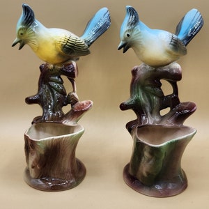 May include: Two ceramic bluebird figurines perched on tree stump planters. The birds are painted with blue, yellow, and white accents. The planters are painted with brown, green, and white accents.