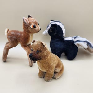 May include: Three vintage plush stuffed animals: a brown deer, a black and white skunk, and a brown dog. The animals are all sitting on a light brown surface.