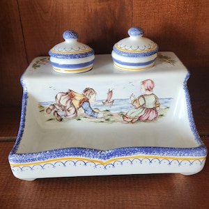 May include: A white ceramic inkwell with two small blue and yellow lidded jars. The inkwell features a hand-painted scene of two children playing by the sea with a sailboat.