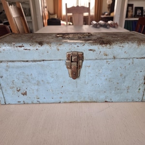 May include: A vintage, rectangular toolbox with a light blue, distressed finish. The metal box features a rusty, metal clasp and handle. The toolbox shows signs of wear and age, with rust spots and faded paint, suggesting it's an antique.