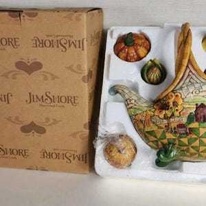 May include: A Jim Shore Harvest Creek decorative cornucopia figurine with a box. The figurine is filled with miniature pumpkins, corn, an apple, and a pear. The cornucopia is painted with a fall harvest scene.
