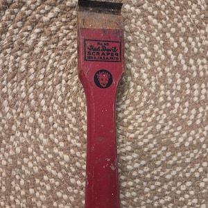 May include: A vintage Red Devil scraper with a red wooden handle and a metal blade. The scraper has the text "Red Devil SCRAPER" and a devil logo. The tool is likely used for removing paint or other materials.