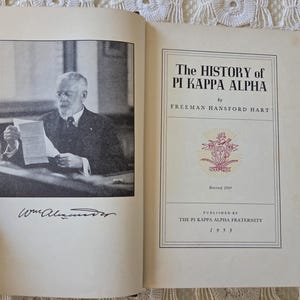 May include: An open book with a black and white photograph of a man in a suit on the left page. The right page displays the title "The History of Pi Kappa Alpha" by Freeman Hansford Hart, published in 1933.