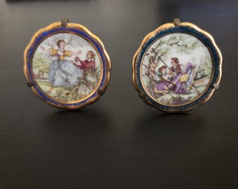 May include: Two antique porcelain miniature plaques. Each plaque features a hand-painted scene of people in a garden setting. The plaques have a scalloped gold rim, a blue border, and a metal loop for hanging. The scenes depict figures in colorful clothing.