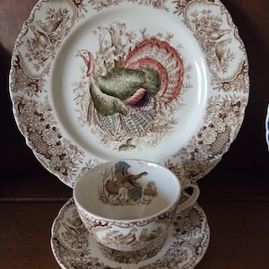 May include: A cream-colored turkey-themed dinnerware set. The set includes a large plate, a teacup, and a saucer. The plate features a detailed illustration of a turkey with a colorful tail. The teacup and saucer have matching turkey and floral designs.