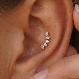 May include: A gold ear piercing stud featuring five small, clear, pear-shaped stones arranged in a curved, fan-like formation. The jewellery is positioned within the conch area of the ear, showcasing a delicate metallic setting and sparkling stones.