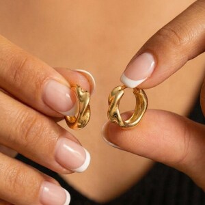 Peut inclure: Une paire de boucles d'oreilles créoles dorées avec un design torsadé unique. Les boucles d'oreilles sont tenues par les mains d'une personne, mettant en valeur leur forme complexe et leur finition polie. Les créoles ont une surface lisse et réfléchissante.