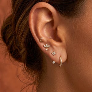 Puede incluir: Primer plano de una oreja adornada con múltiples pendientes dorados. Los pendientes incluyen una media luna, un diseño floral, un pequeño aro y varios pendientes de botón, todos adornados con pequeñas piedras brillantes.