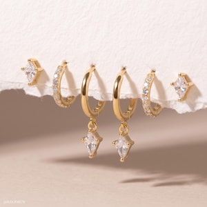 May include: A set of six gold-toned earrings including two kite-shaped studs with clear stones, two small pavé-set hoop earrings, and two hoop earrings with dangling kite-shaped charms featuring clear stones. The text @ALEXJEW978 is visible.