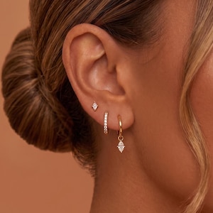 Può includere: Un set di tre orecchini color oro composto da un piccolo perno con taglio a marquise, un sottile orecchino a cerchio incastonato con piccole pietre trasparenti e un cerchio dorato con un ciondolo a forma di pietra trasparente.