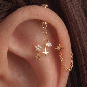 Könnte beinhalten: Ein goldfarbenes Ohrpiercing-Schmuckset mit mehreren hängenden Elementen. Das Design umfasst einen mit klaren Steinen besetzten Sternanhänger, einen schlichten goldenen Stern, einen runden klaren Stein und einen vierzackigen Stern, die durch feine Ketten verbunden sind.