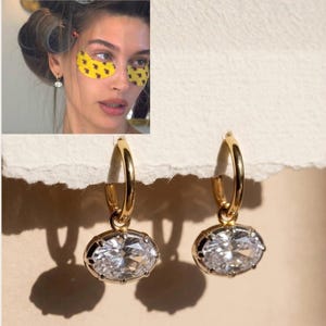 May include: A pair of gold hoop earrings featuring dangling oval-shaped clear faceted stones. Each earring consists of a polished gold hoop connected to a bezel-set oval crystal that catches the light, creating a bright and reflective appearance.