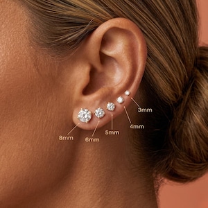 May include: An ear adorned with six diamond stud earrings in sizes 3mm, 4mm, 5mm, 6mm, and 8mm. The earrings are set in gold. The ear is set against a peach-colored backdrop.