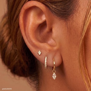 Op de afbeelding: Een set van drie gouden oorbellen: een kleine marquise-vormige knop, een dunne ring bezet met kleine doorzichtige steentjes en een eenvoudige gouden ring met een bungelende marquise-vormige hanger in goudkleurig metaal.