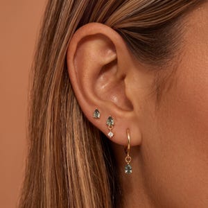 Può includere: Un set di tre orecchini in oro indossati su un solo orecchio. L'orecchino superiore è un piccolo perno con pietra preziosa verde a forma di pera. L'orecchino centrale è una goccia di pietra verde con una piccola pietra chiara. L'orecchino inferiore è un cerchio d'oro con un ciondolo in pietra verde a forma di pera.