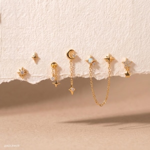 May include: A collection of gold-tone earrings featuring celestial motifs including stars, a crescent moon, and a small opal-like stone. The set includes individual studs and a double-stud earring connected by a delicate gold chain. Text reads @ALEXJEWS78.