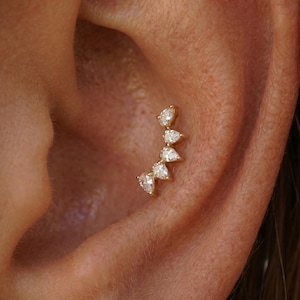 Pode incluir: Um piercing de orelha em ouro com uma fila curva de cinco pedras preciosas transparentes em forma de pera, dispostas em leque. A joia está montada numa base de metal de ouro amarelo e posicionada na cartilagem da orelha humana.