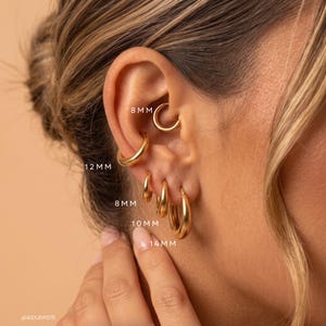 Può includere: Una collezione di orecchini a cerchio color oro in varie dimensioni, etichettati 8mm, 10mm, 12mm e 14mm. Gli orecchini sono indossati in molteplici fori lungo l'orecchio, caratterizzati da una finitura liscia e lucida e un design circolare.