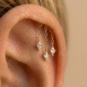 Puede incluir: Pendientes de cadena dorada con tres colgantes en forma de diamante. Los pendientes están sujetos a la oreja, mostrando el delicado diseño y el brillo de las piedras preciosas. La joya se encuentra sobre el fondo de la oreja.