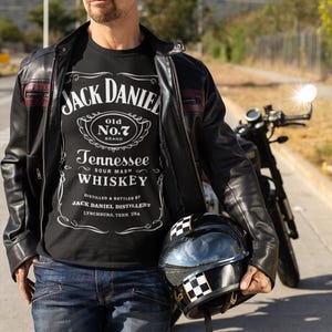 Puede incluir: Un hombre con una chaqueta de cuero negra sobre una camiseta negra con el logo de "Jack Daniel's". Sostiene un casco de moto negro con un diseño de cuadros. Se ve una moto al fondo.