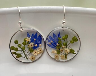 May include: A pair of circular drop earrings featuring clear resin encasing pressed botanical elements. Each earring contains vibrant blue petals, small white flowers, and green leafy stems, suspended from silver-toned metal ear wires.