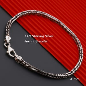 May include: A 925 sterling silver foxtail bracelet with a lobster claw clasp. The metal link chain features a woven design and is marked with 925 and Italy on the clasp. The text 925 Sterling Silver Foxtail Bracelet and 3 mm are visible.