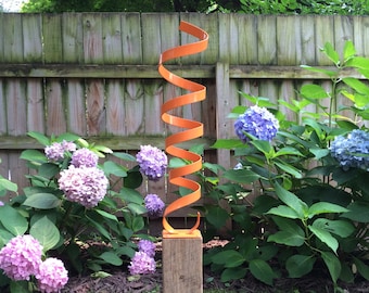 Op de afbeelding: Oranje metalen sculptuur in een spiraalvorm gemonteerd op een houten paal in een tuinomgeving. De sculptuur is omringd door roze hortensia's.