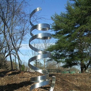May include: A modern, abstract sculpture made of polished stainless steel. The sculpture is a spiral shape and is mounted on a square base. The sculpture is outdoors on a rocky surface.